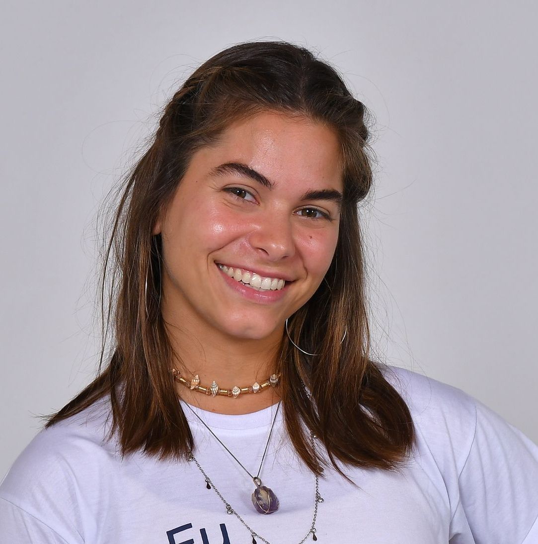 Jovem mulher de cabelos castanhos sorri largamente, vestindo uma camisa branca, colares e brincos de argola.