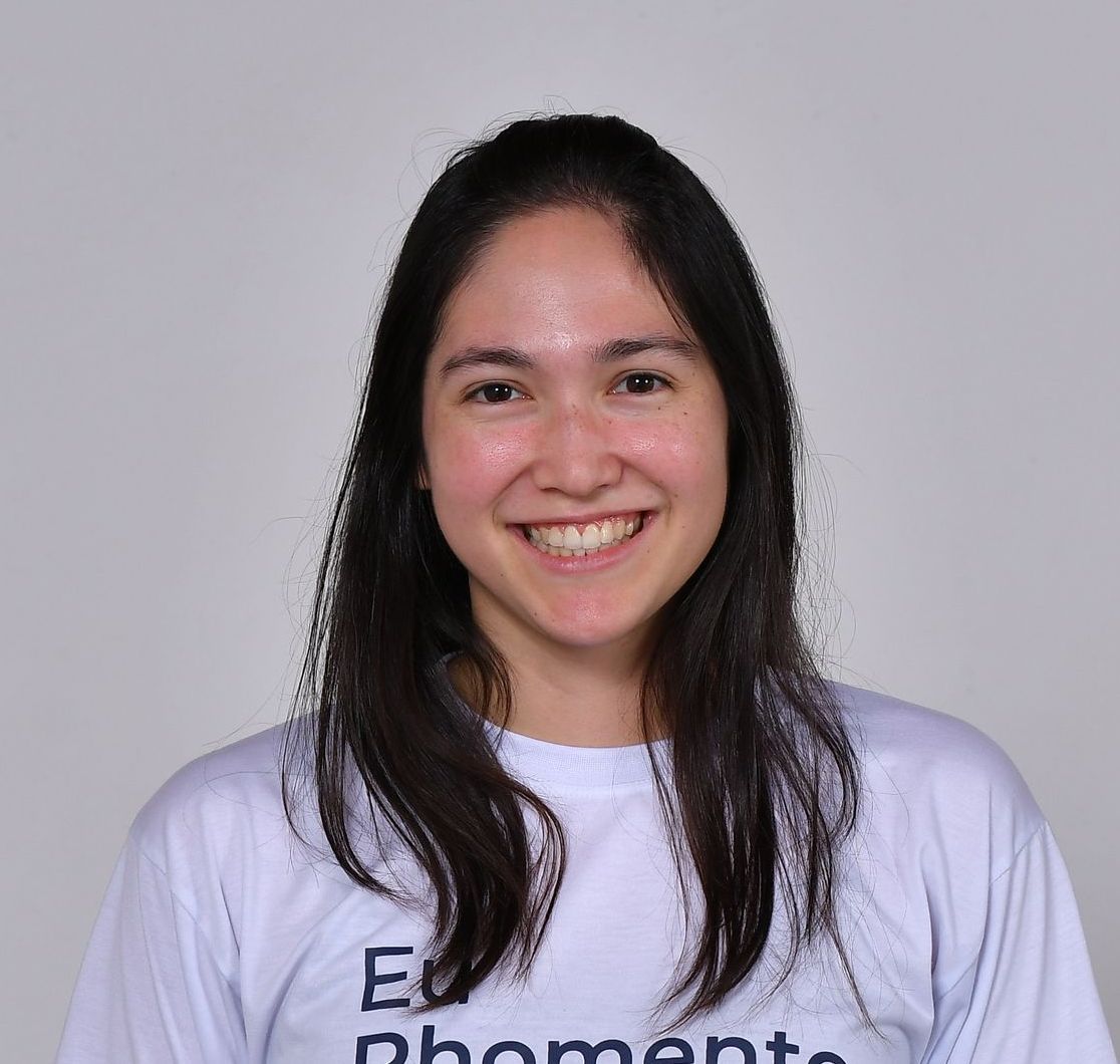 Mulher de cabelos escuros, sorrindo, vestindo uma camisa branca com texto, sobre um fundo branco.