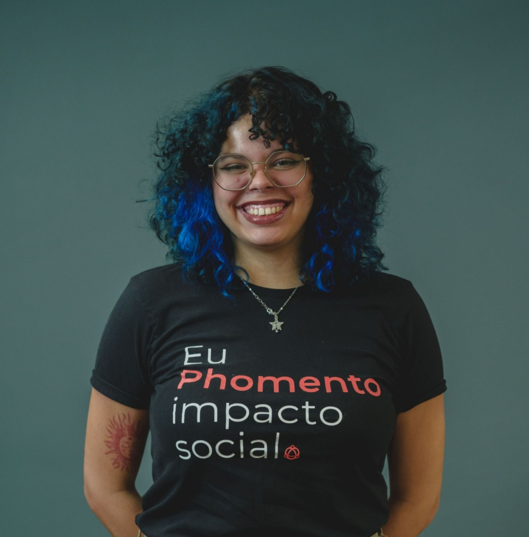 Uma pessoa sorridente com cabelo encaracolado com reflexos azulados, vestindo uma camiseta preta com a inscrição 