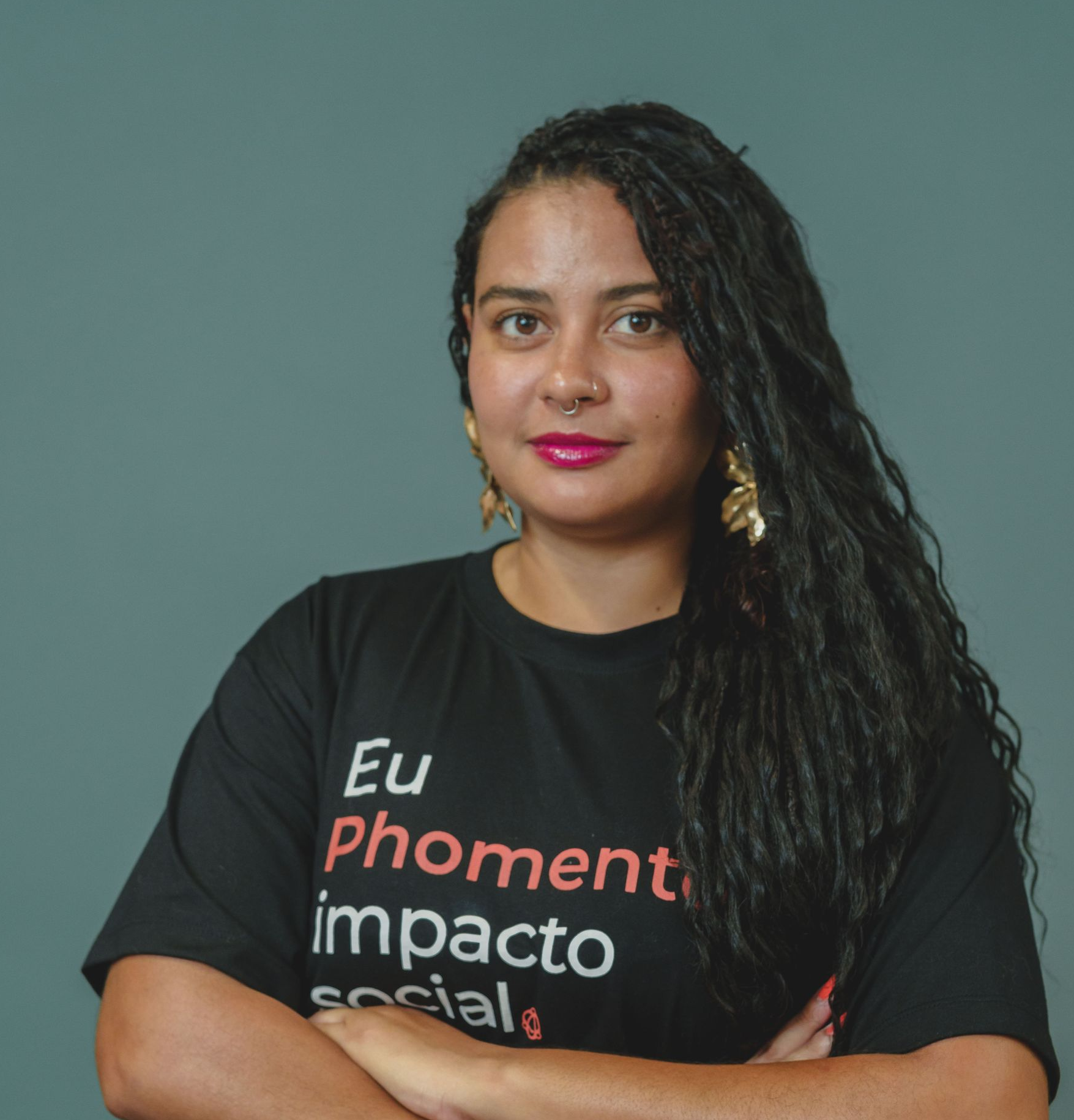 Mulher com tranças escuras, vestindo uma camiseta branca, brincos grandes e um piercing no nariz, sorrindo.