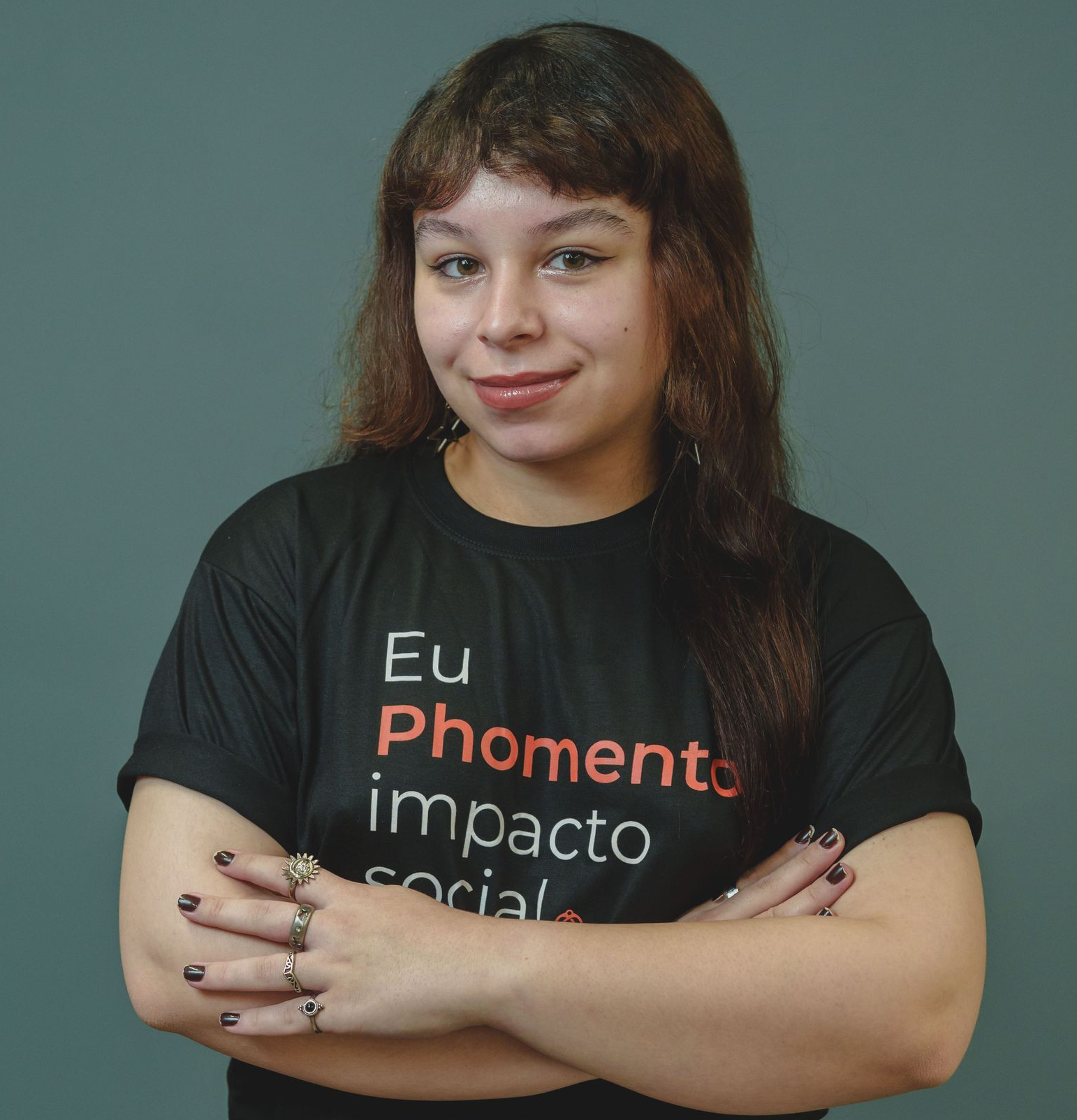 Uma pessoa sorridente, de cabelos longos e escuros com franja, vestindo uma camisa branca, está em pé em frente a uma porta de madeira e uma faixa colorida.