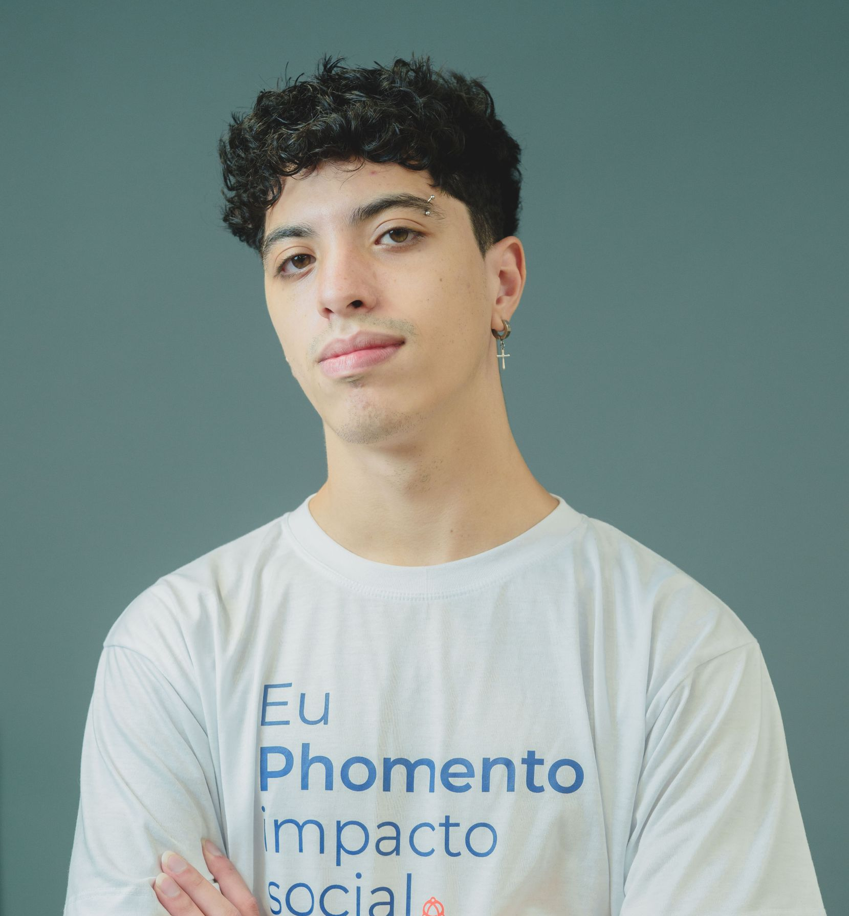 Jovem de cabelos escuros e cacheados, com uma sobrancelha perfurada, sorri e veste uma camiseta branca.