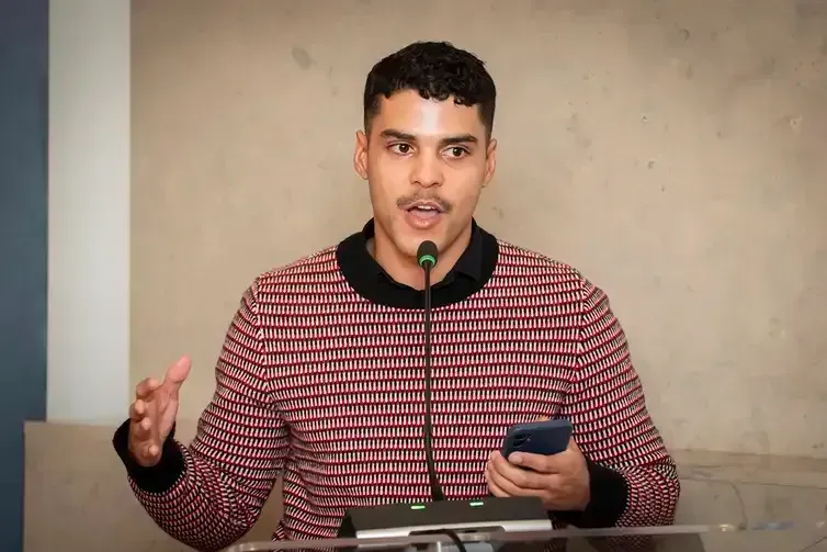 Homem com cabelos escuros e bigode falando em um microfone, gesticulando com as mãos enquanto segura um telefone.