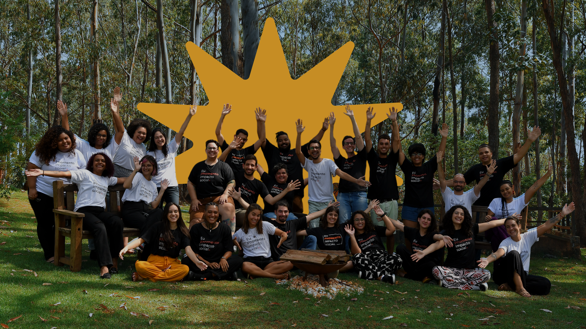 Um grande grupo de pessoas sorrindo e acenando enquanto posam para uma foto em um gramado em uma área arborizada.