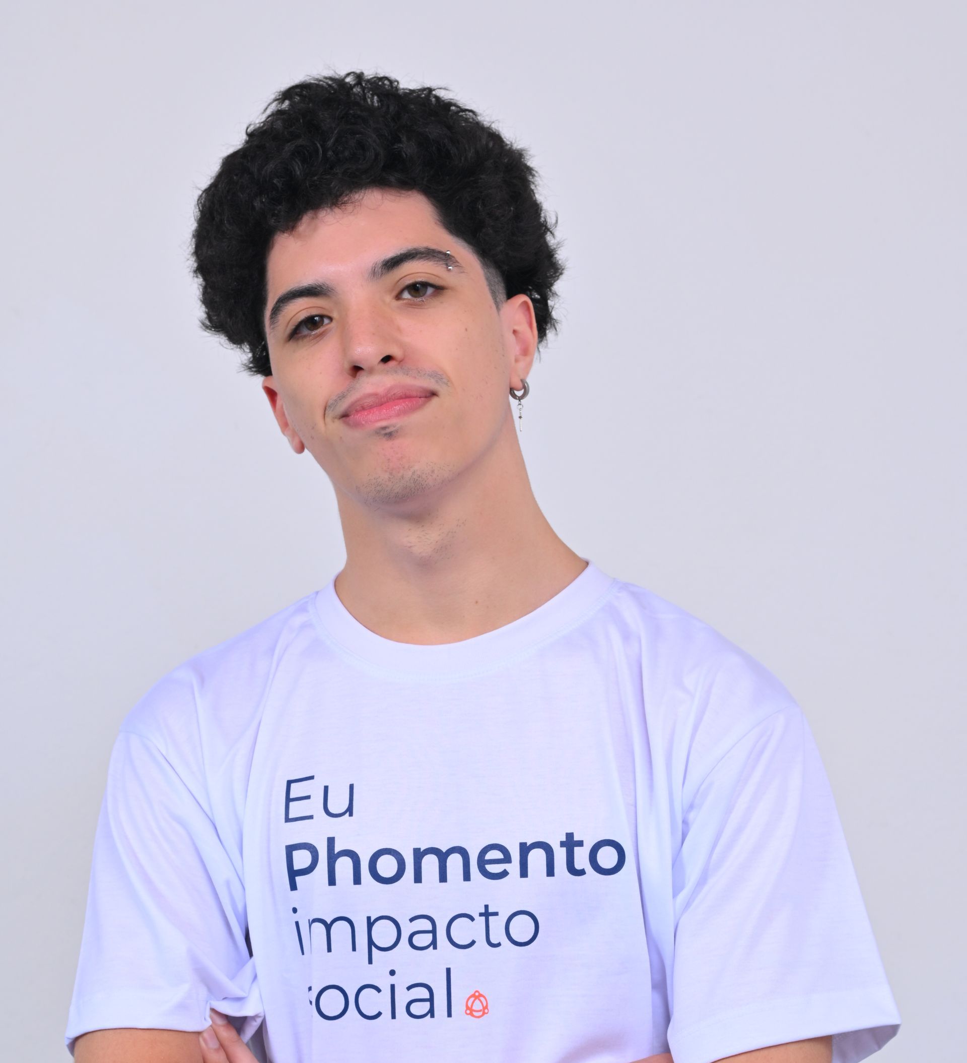 Jovem de cabelos escuros e cacheados, com uma sobrancelha perfurada, sorri e veste uma camiseta branca.