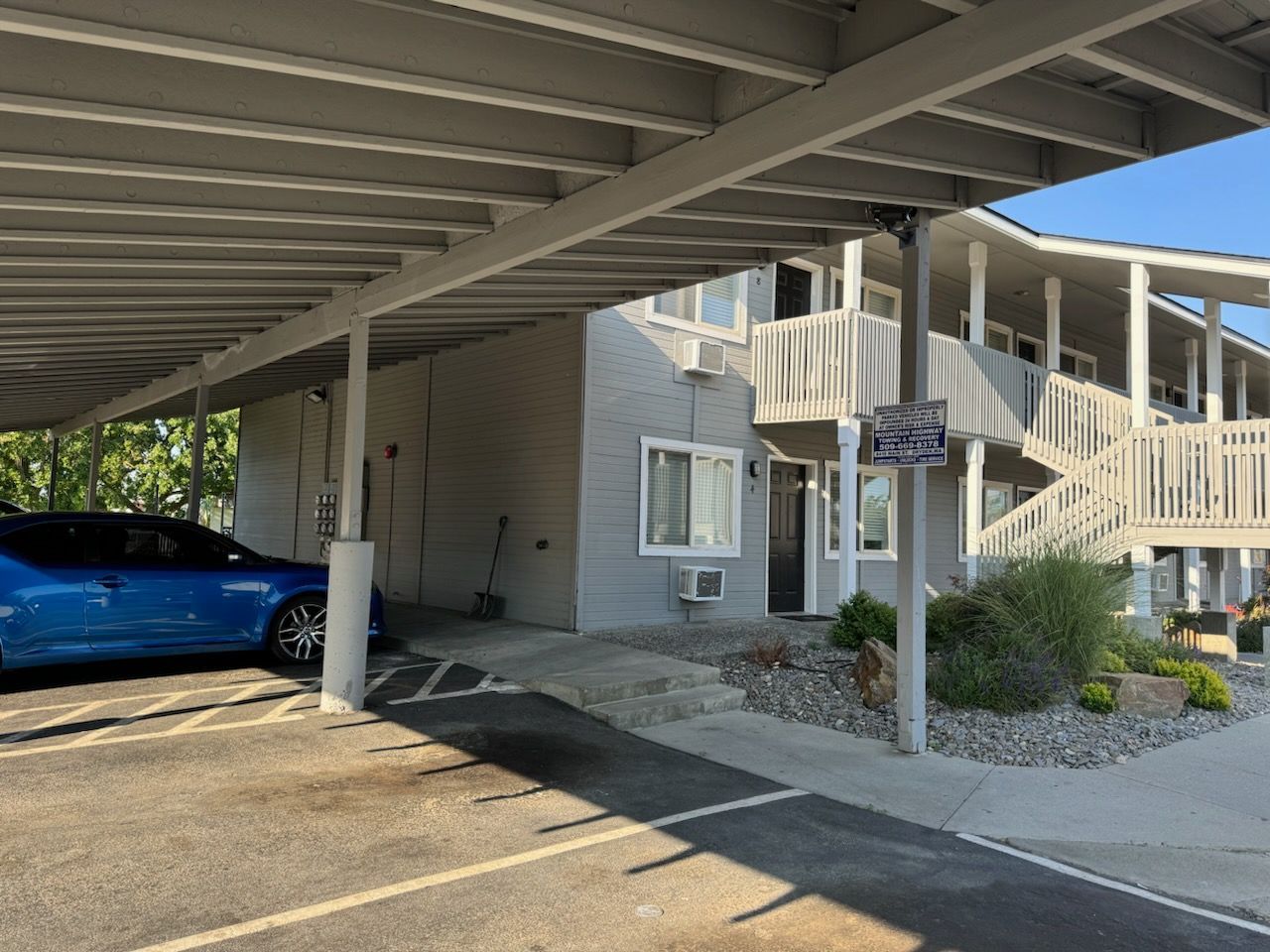 A blue car is parked under a covered parking lot in front of a building.