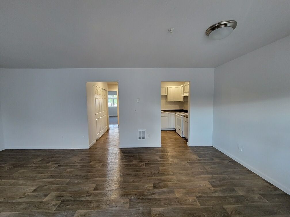 An empty living room with hardwood floors and white walls leading to a kitchen.