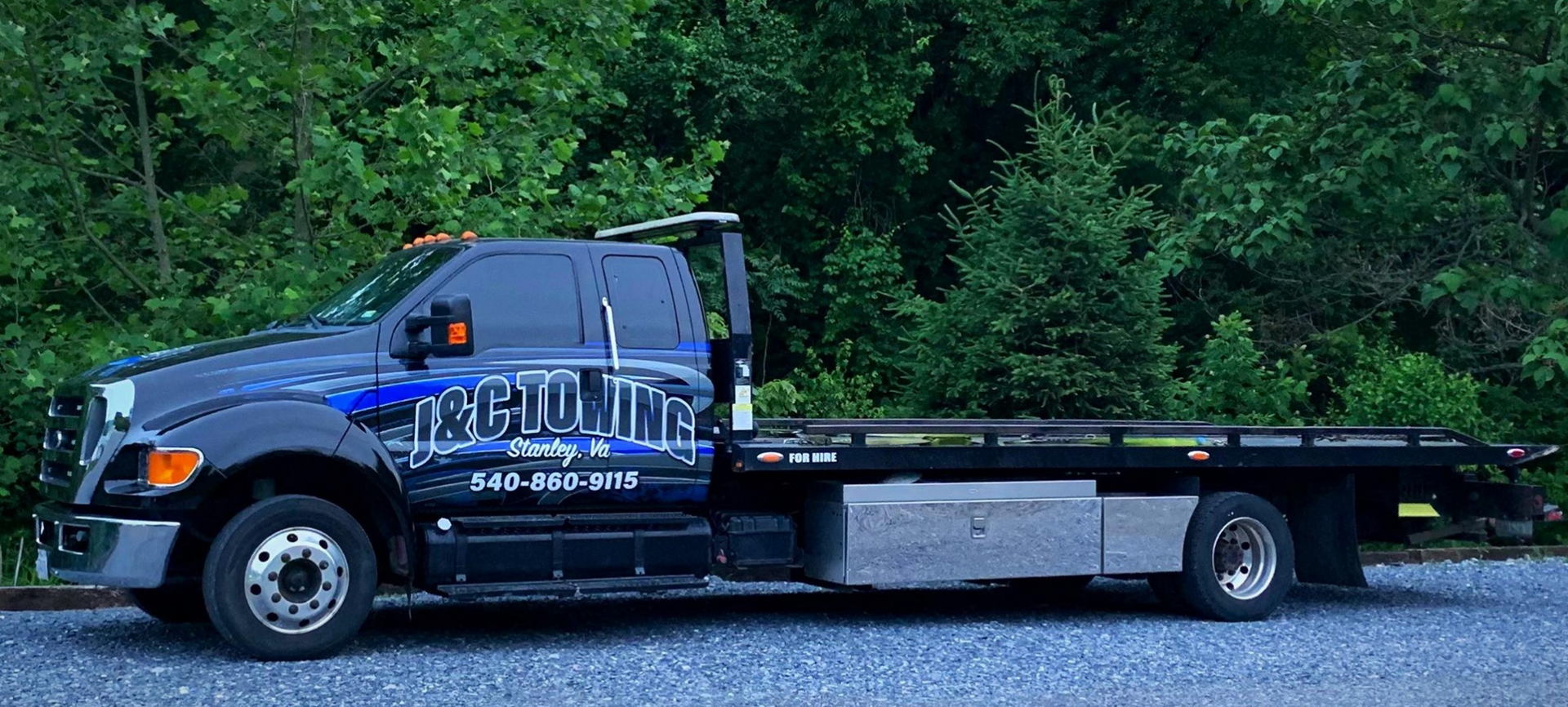 Black tow truck with blue logo parked on gravel in front of green foliage.
