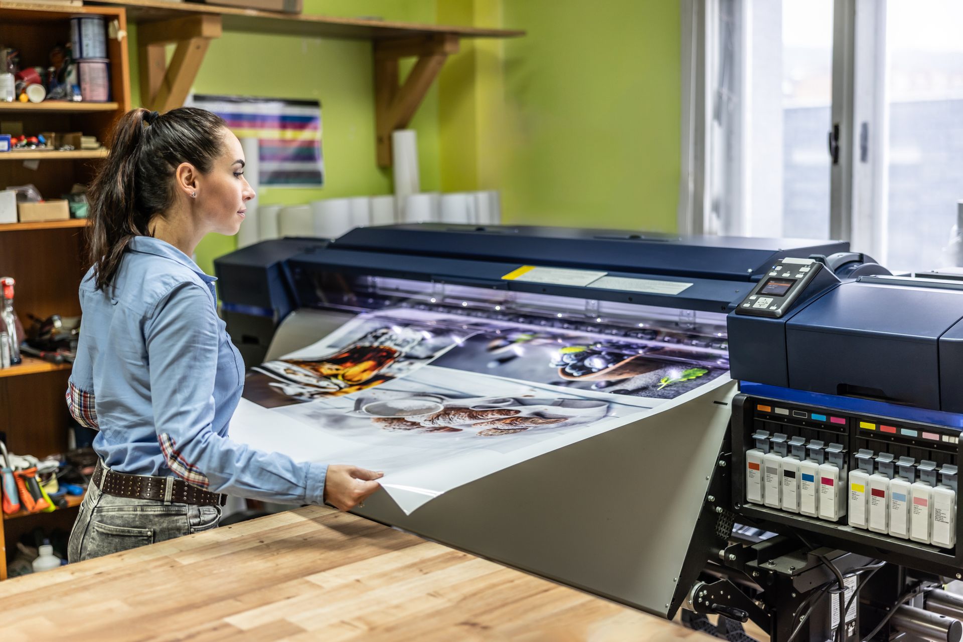 Person handles vibrant prints from large digital printer in a busy, well-stocked print shop.