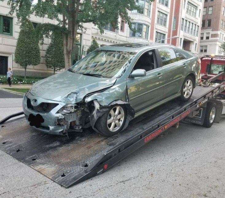 Green tow truck carrying a black car on a city street with blurred background.