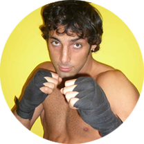 A shirtless man wearing black boxing gloves stands in front of a yellow wall