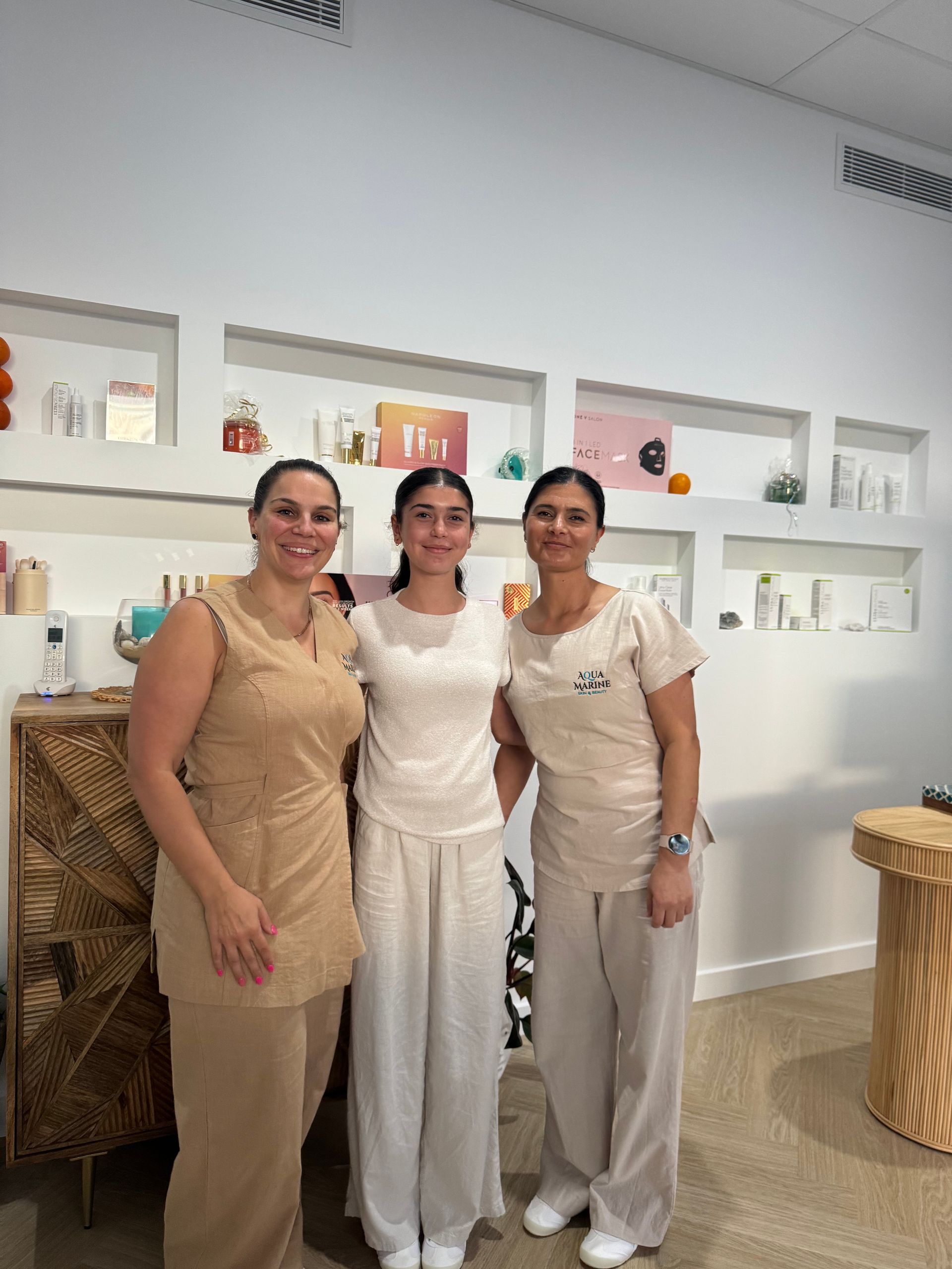 Three people in cream-colored outfits pose in a store. Shelves display beauty products — Aqua Marine Skin and Beauty in Tuncurry, NSW