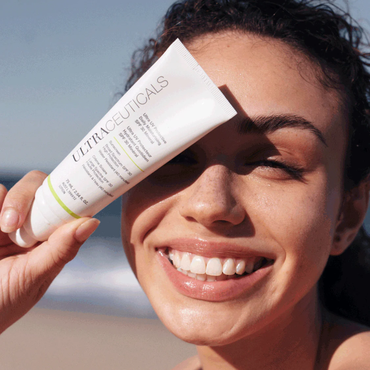 Woman smiling, holding Ultraceuticals sunscreen on beach, shielding one eye. — Aqua Marine Skin and Beauty in Tuncurry, NSW