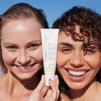 Two women smiling, holding a white tube of sunscreen under a bright blue sky. — Aqua Marine Skin and Beauty in Tuncurry, NSW