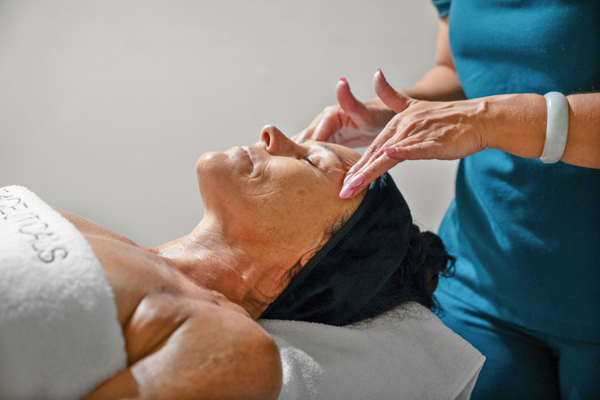 Woman receiving a facial in a spa; masseuse's hands on her face, teal uniform. — Aqua Marine Skin and Beauty in Tuncurry, NSW