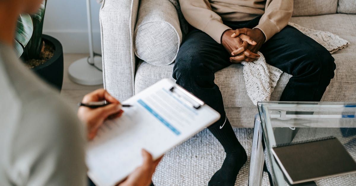 A man is sitting on a couch while a woman holds a clipboard for therapy