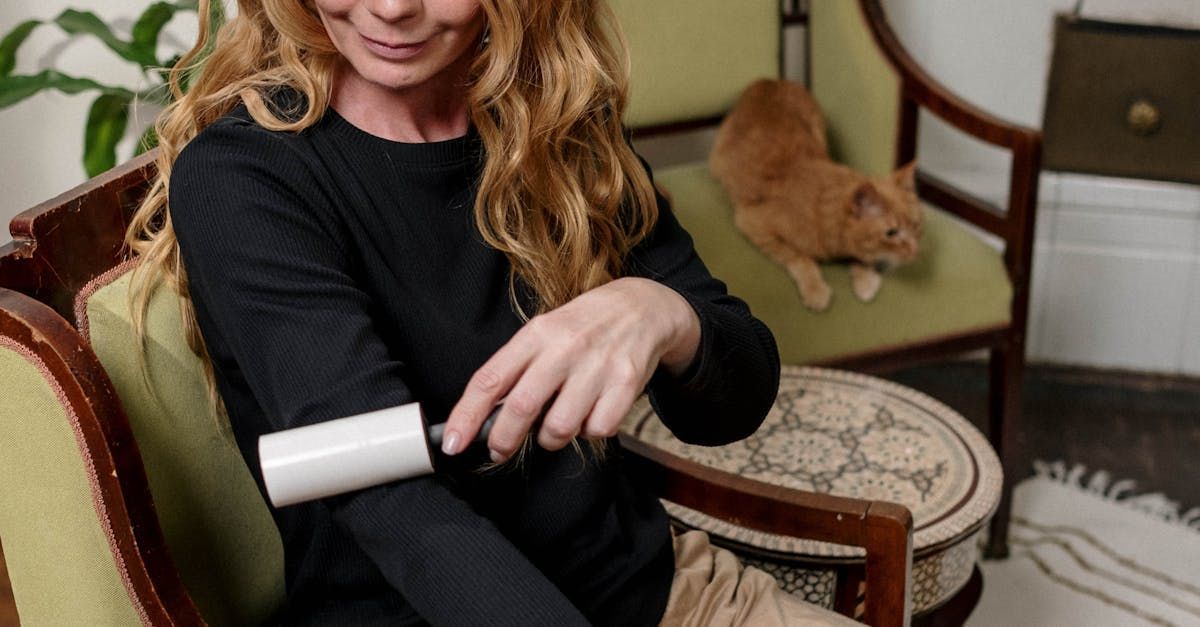 Woman using lint roller on her arm while an orange cat sits on a chair, likely removing cat hair.