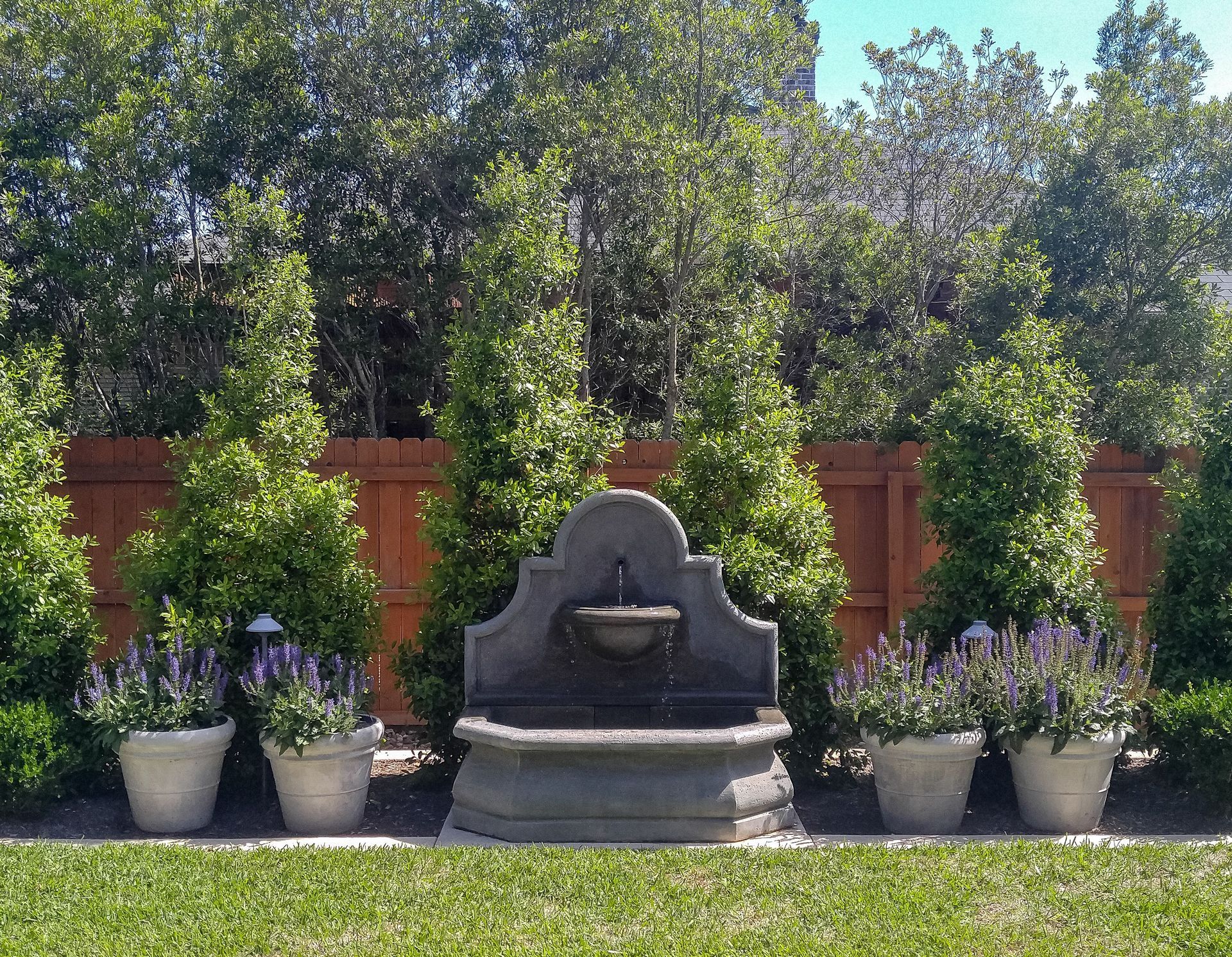 A fountain is surrounded by potted plants and a wooden fence