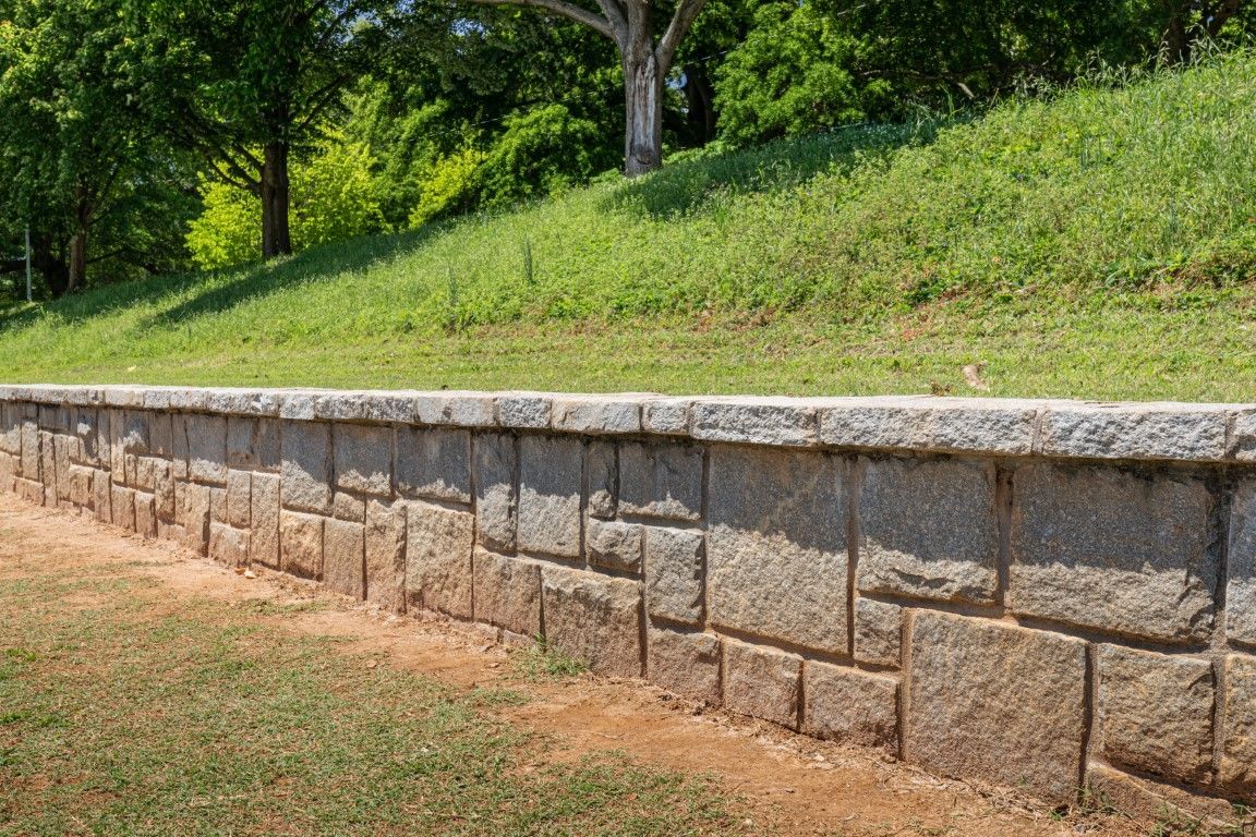 Stone retaining wall with a grassy slope and trees in the background.