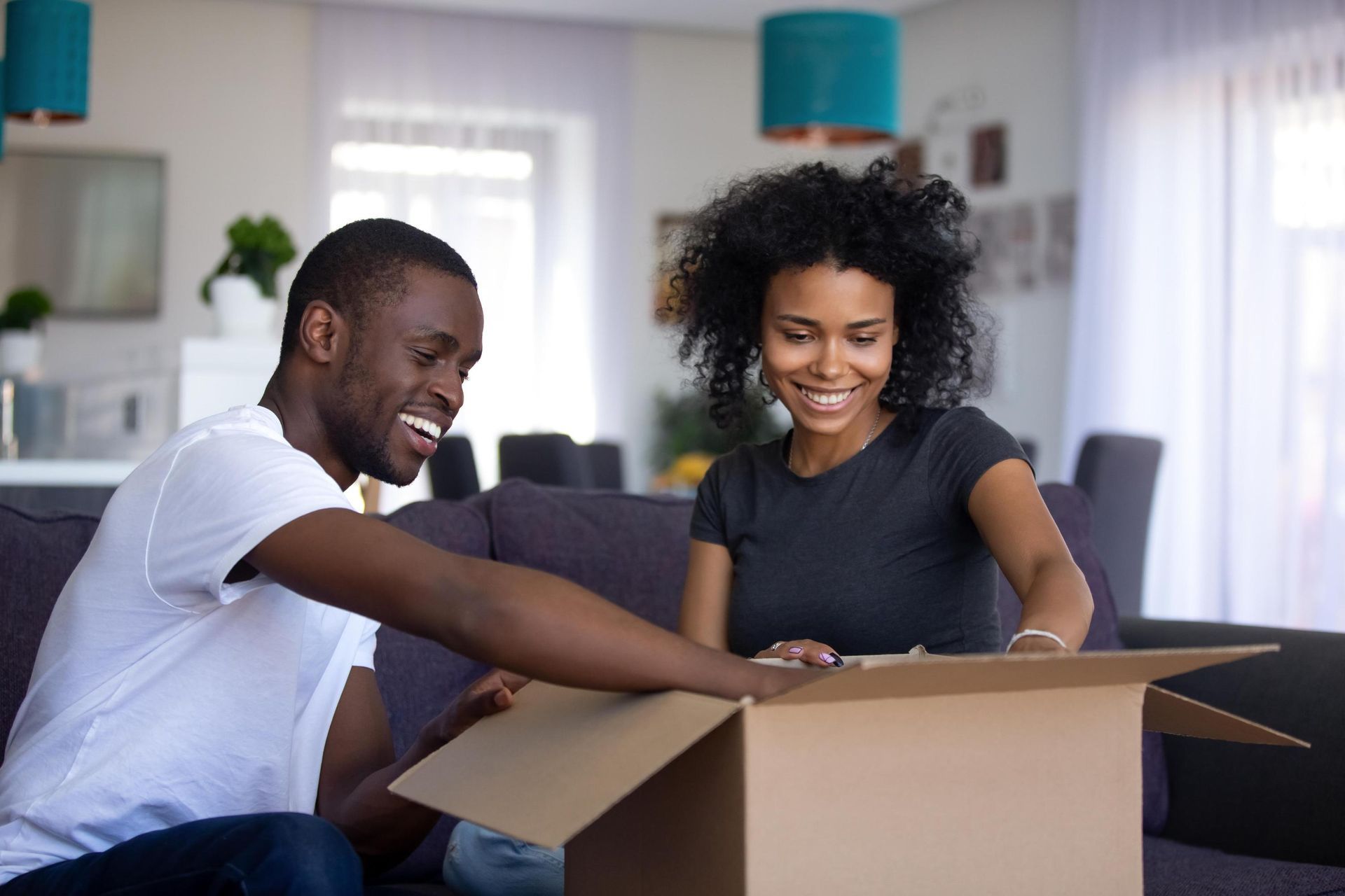 A smiling Black couple unpacks a cardboard box on a sofa in their home.