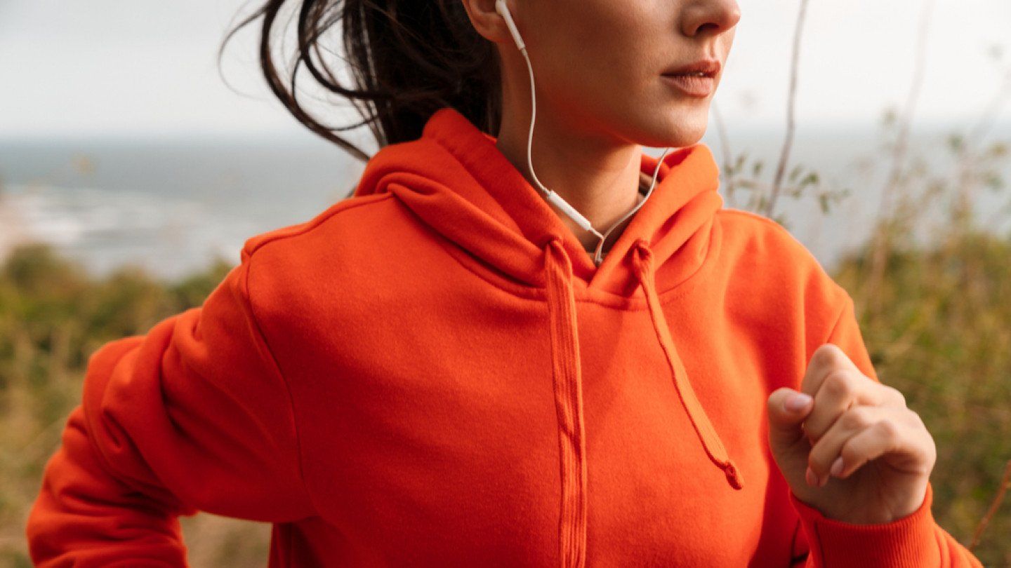 A woman in an orange hoodie is running with headphones on.