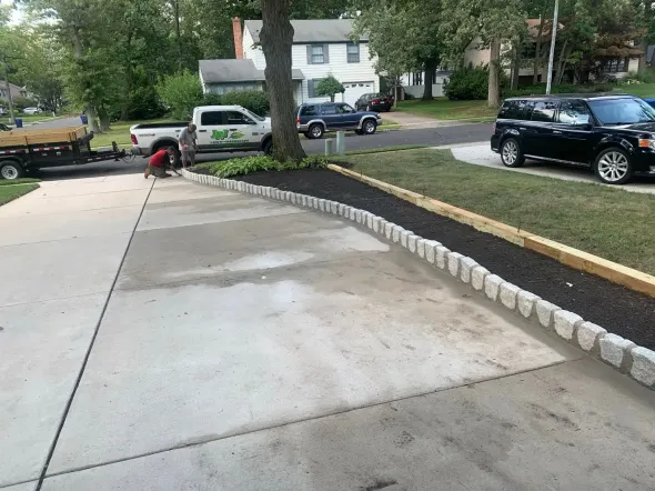 Workers landscaping a driveway, placing edging stones and mulch, with trucks and homes in the background.