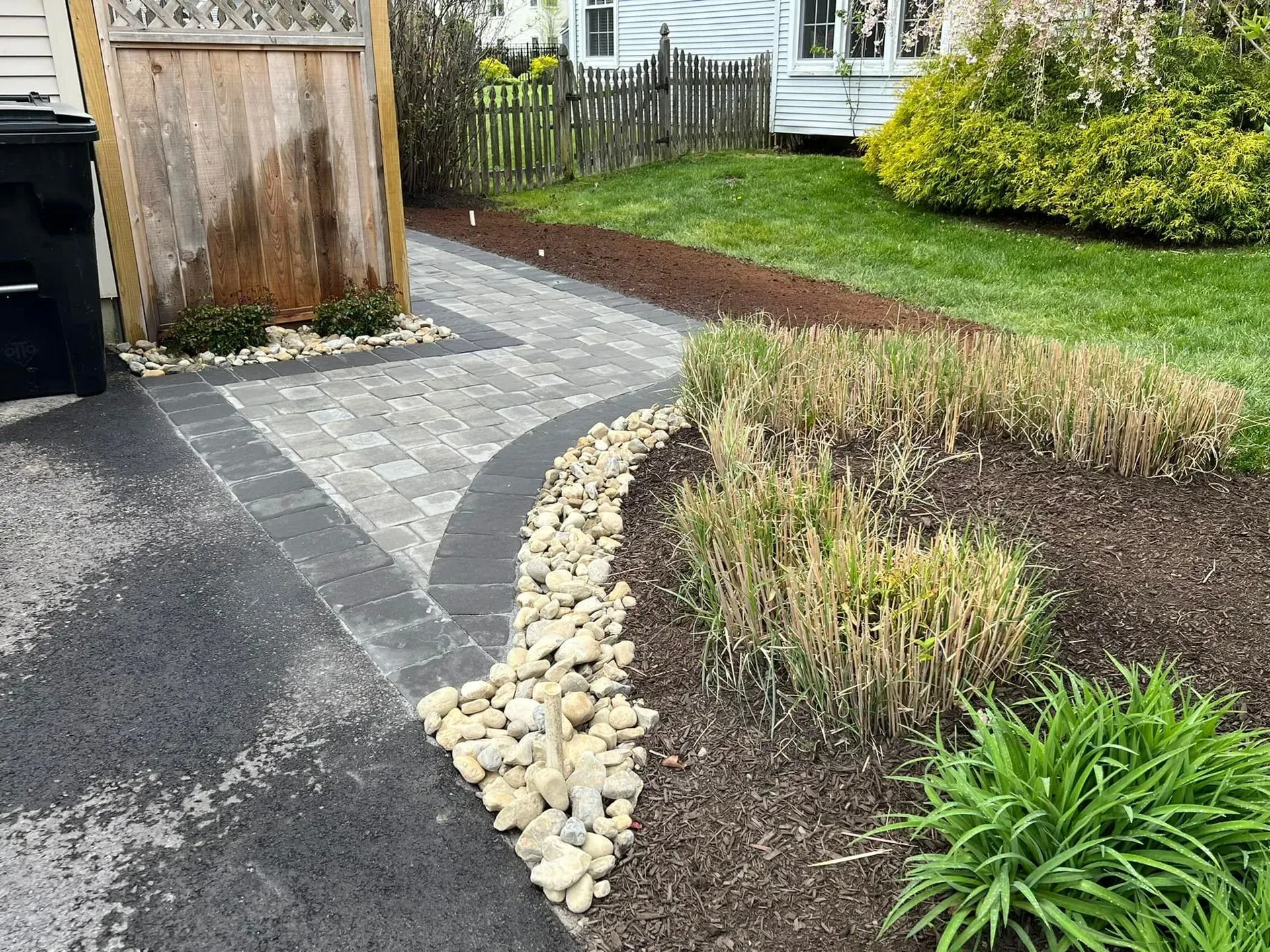 A paved walkway bordered by landscaping with rocks and mulch leads to a house with a green lawn.