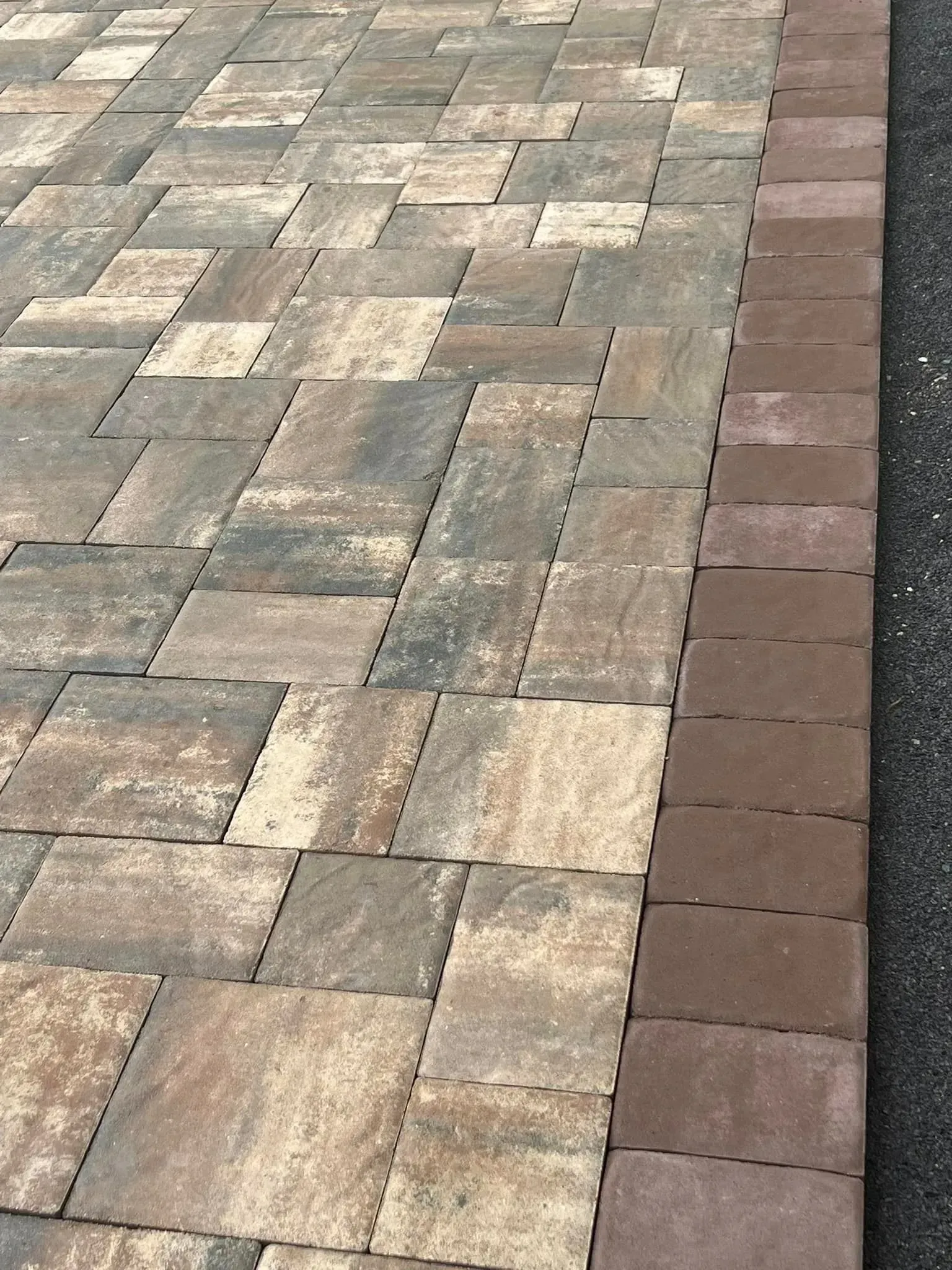 Paver patio with a dark border. Brown and tan pavers are laid in a random pattern next to a black surface.