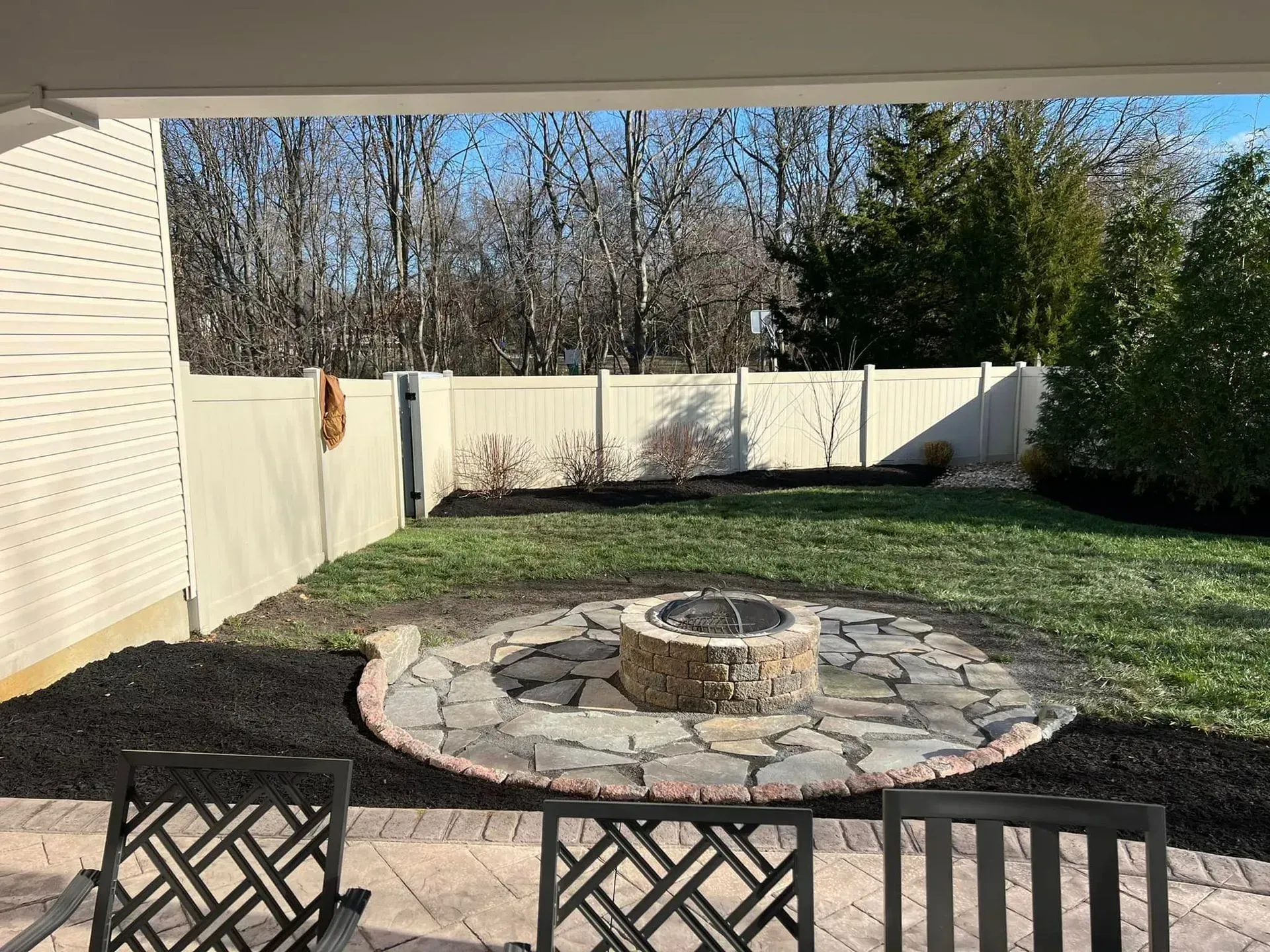Backyard with fire pit, surrounded by stone and mulch, green grass, and white fence.