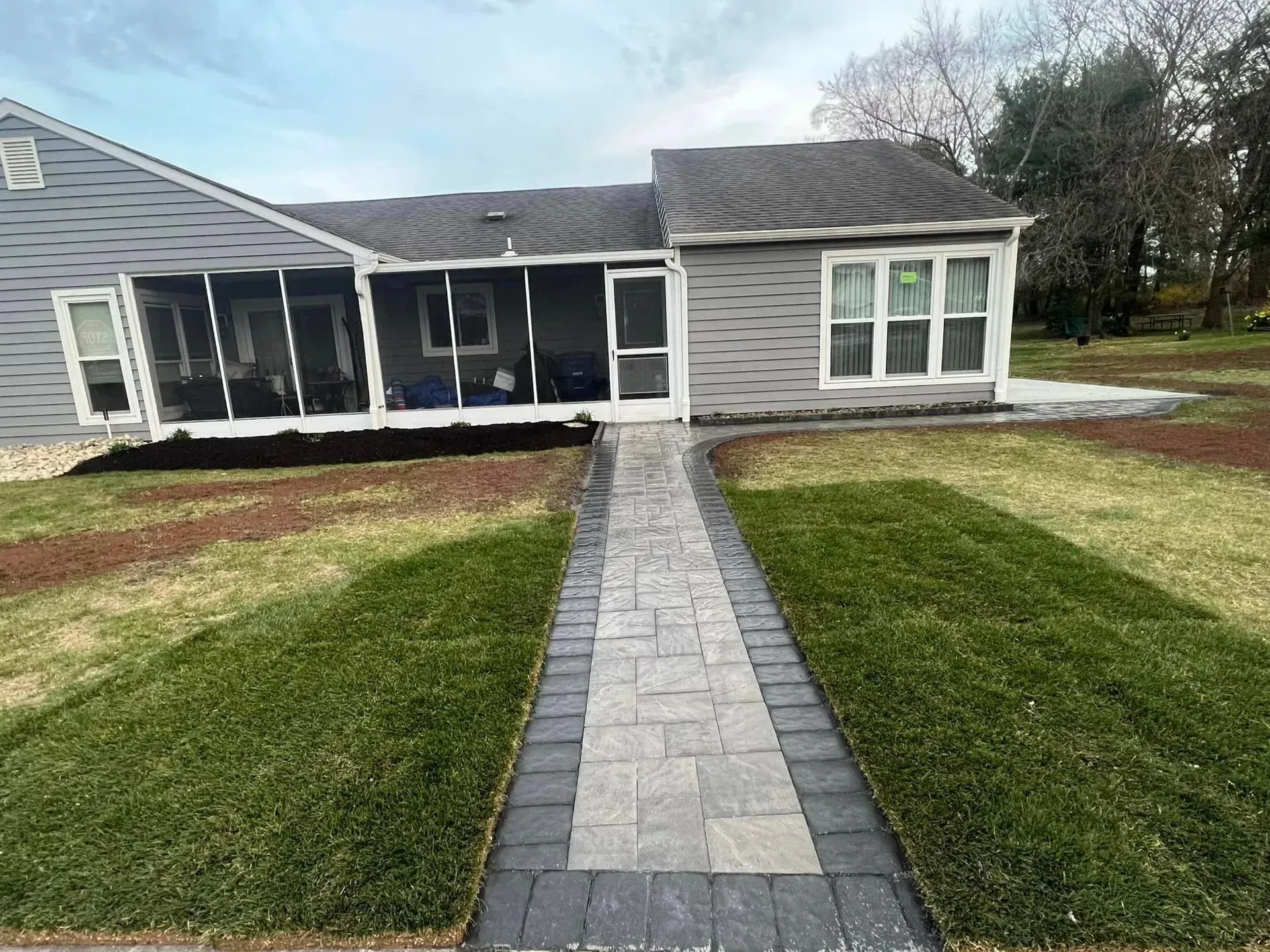 Stone walkway leads to a screened porch on a gray house, flanked by green grass.