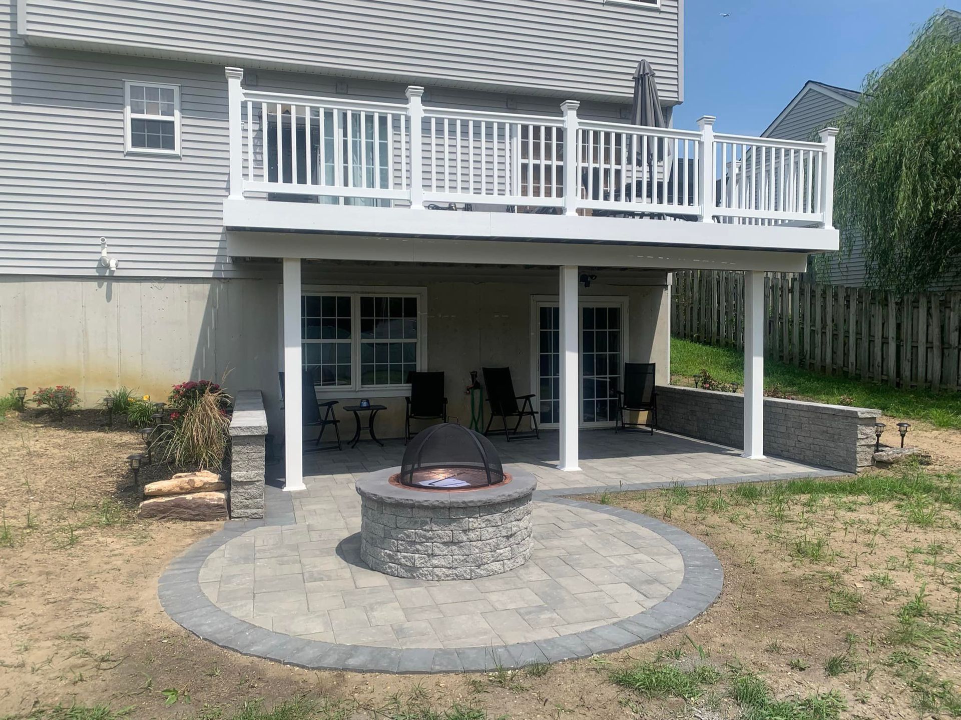 A patio with a fire pit under a white deck. Gray pavers, chairs, and a house in the background.