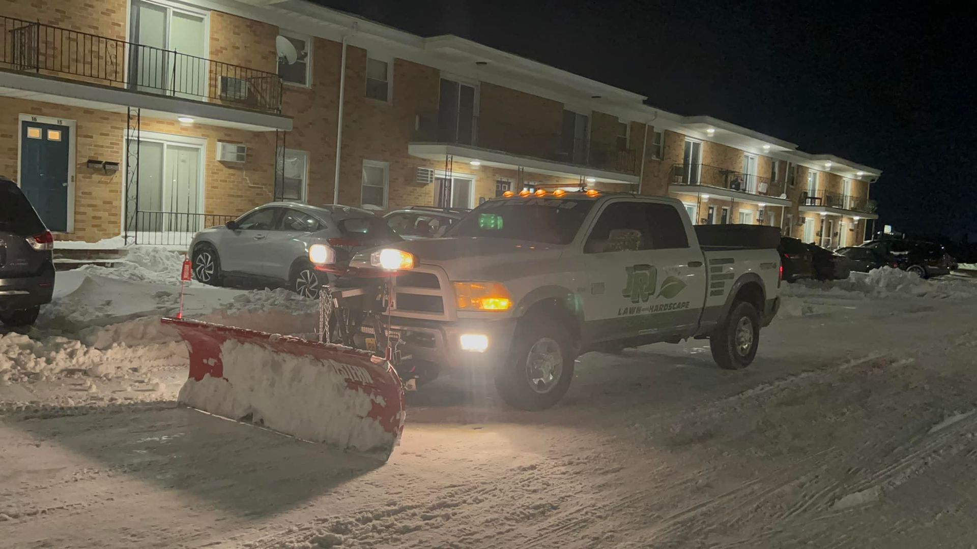 Snowplow clearing a snow-covered street in front of a brick apartment building at night.