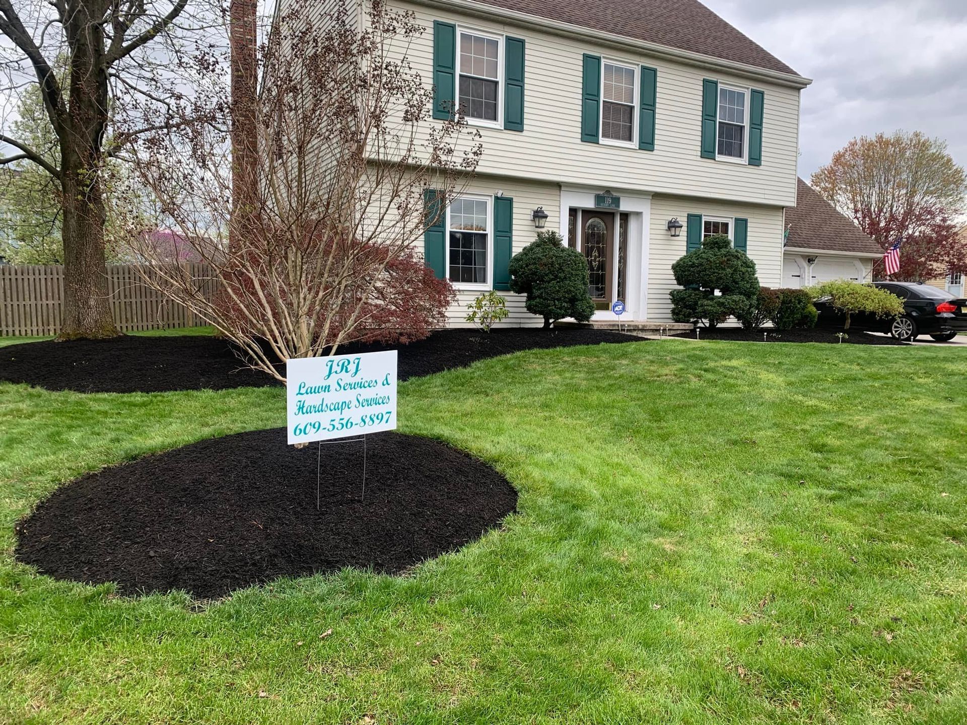 Two-story house with green shutters, black mulch, and a lawn sign advertising lawn and roofing services.