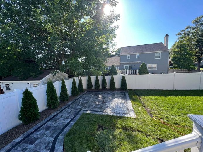 Backyard with a gray paved patio, white fence, evergreen trees, and a gray house.