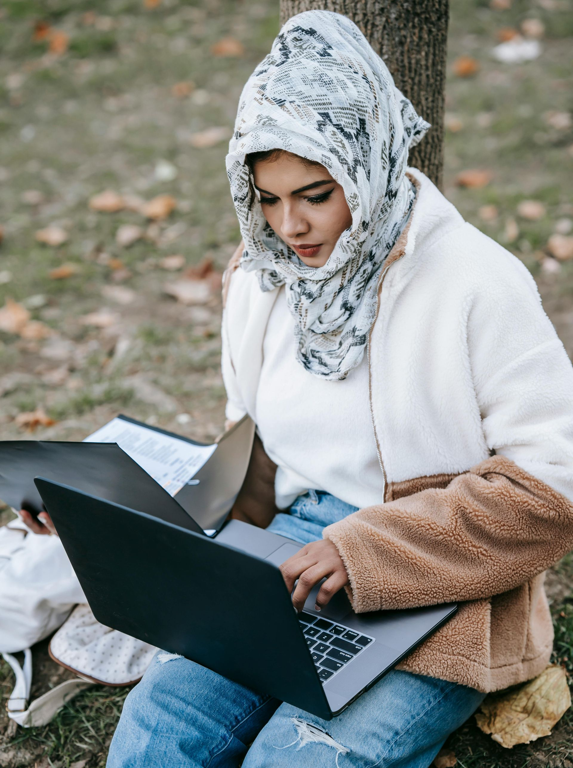 A woman is sitting under a tree using a laptop computer.