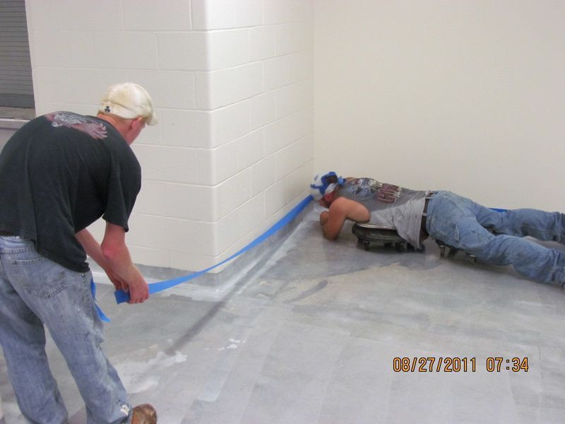 Two men applying blue tape to a floor corner in a room with white walls, possibly for painting or sealing.