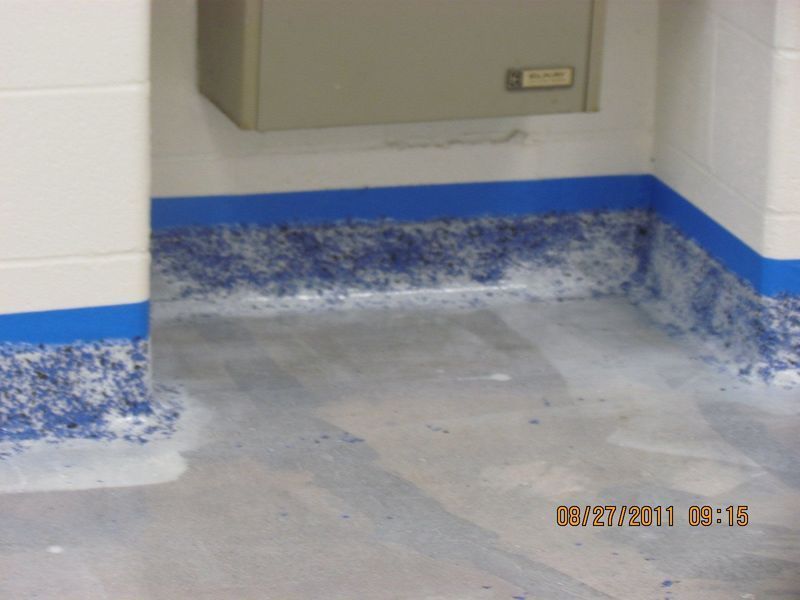 Blue painter's tape lines walls; floor has blue specks in coating. White walls, grey floor in a room.