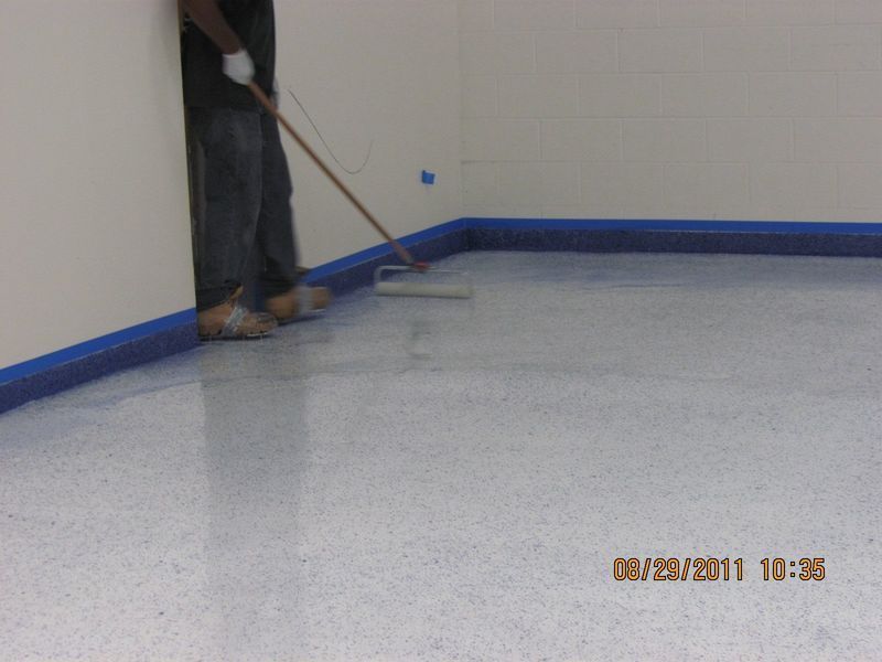 Person applying coating to a speckled floor with a roller. Blue tape protects the walls.