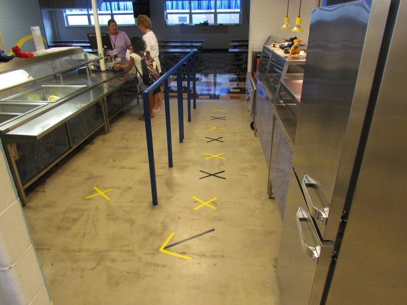 People at a food counter with floor markers indicating distancing. Yellow Xs and an arrow on the floor.