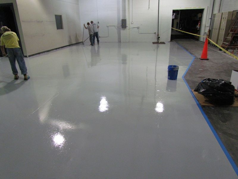 Workers applying a light gray epoxy coating to a large warehouse floor.