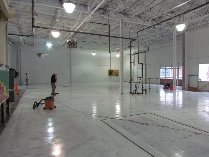 Large, empty, white-walled room with shiny floor. A person vacuums; pipes run along walls and ceiling.