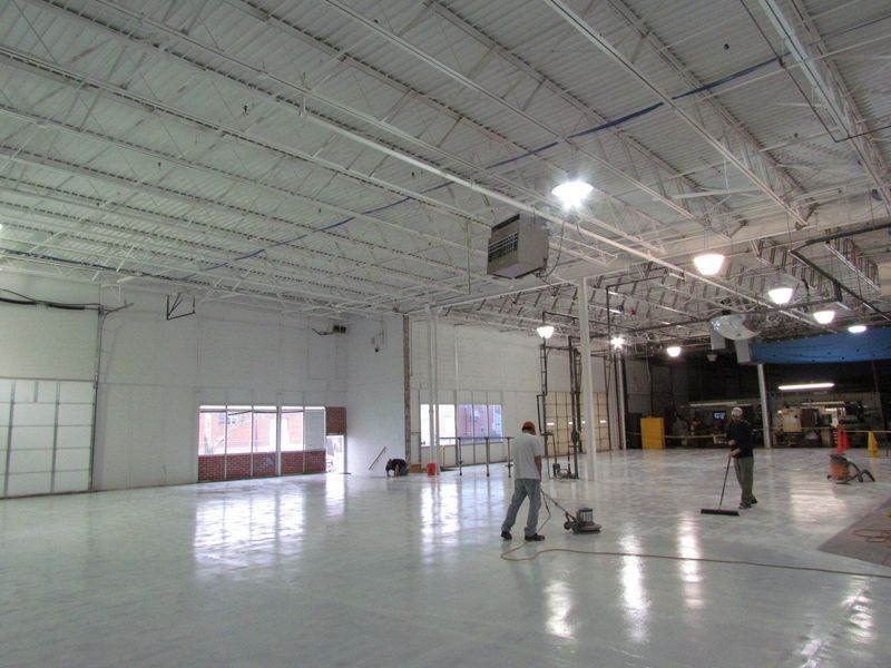 Two workers cleaning a large, white-floored warehouse. One uses a floor machine, the other a broom.