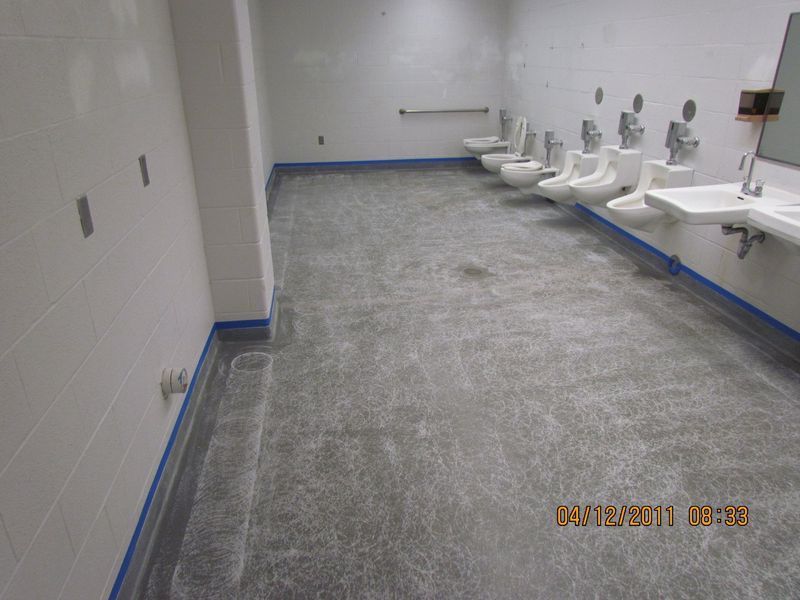 Public restroom with gray concrete floor, white walls, toilets, urinals, sinks. Blue tape along the edges.