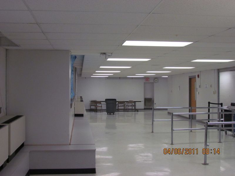 Empty, white-walled room with tables and chairs in the background; metal railings in the foreground; fluorescent lights.