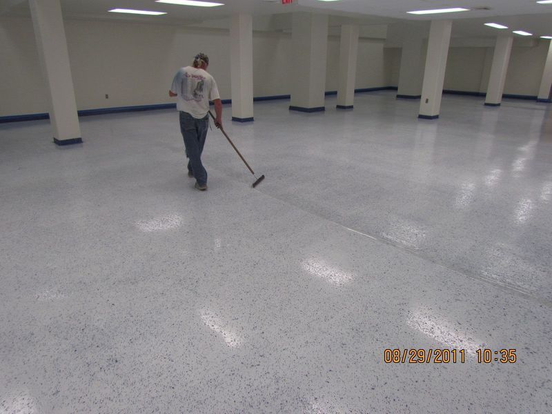 Person sweeping a large, light-colored, speckled floor in an empty room with white pillars and trim.