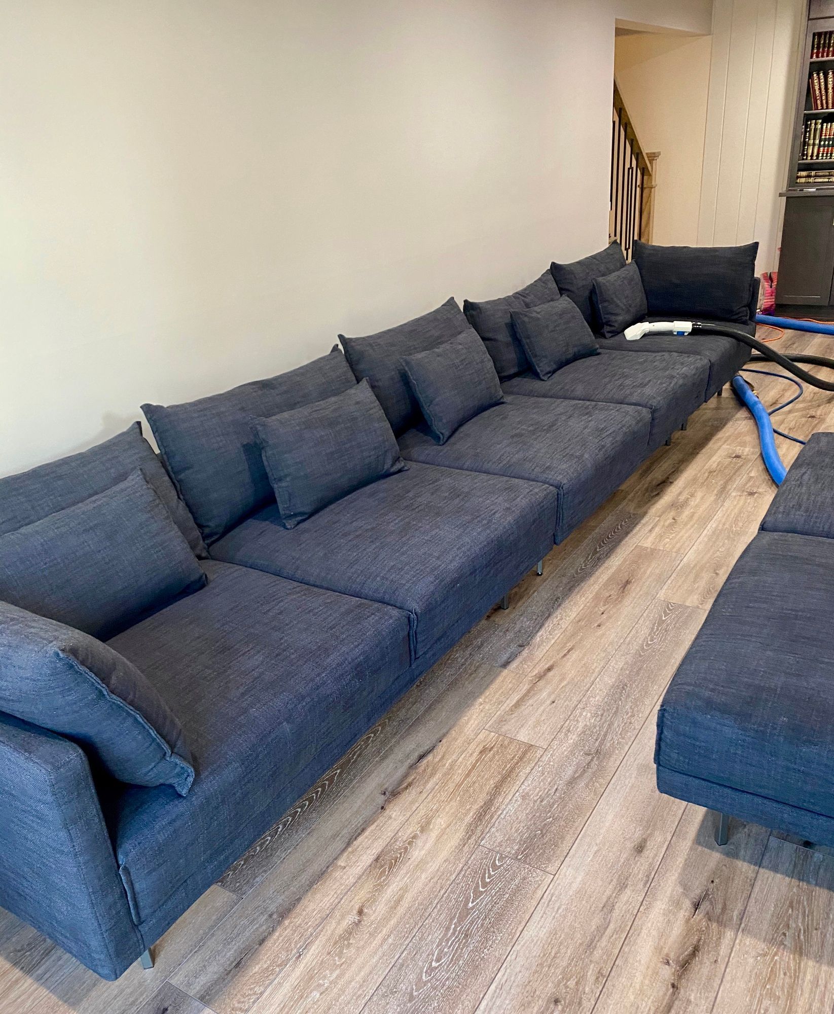 A living room with a couch , ottoman , and vacuum cleaner.