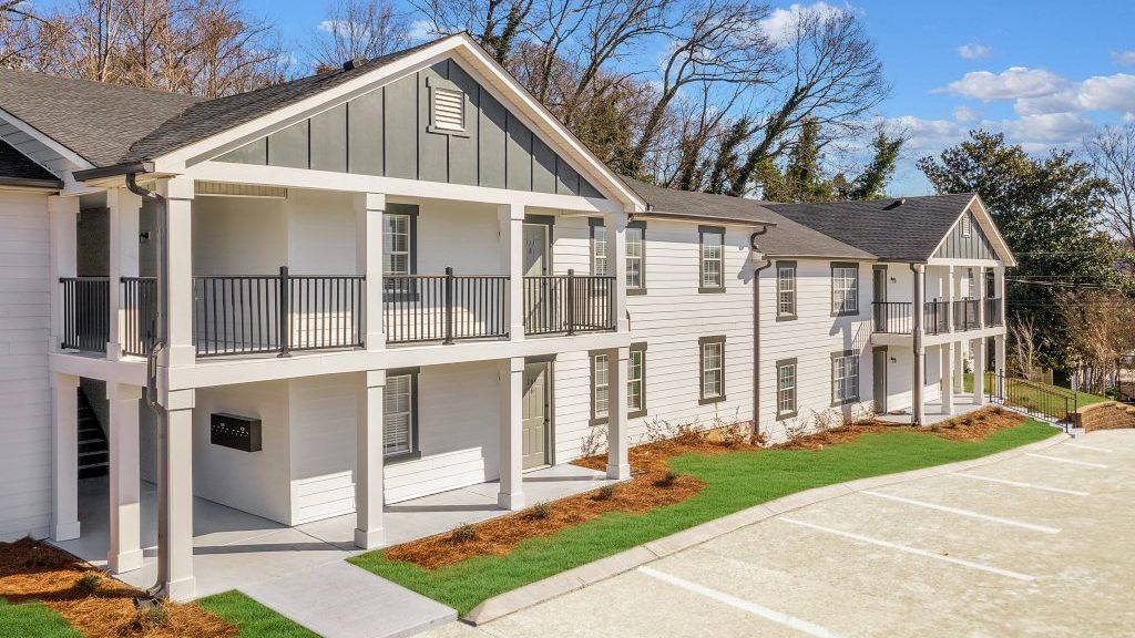 A white apartment building with a balcony and a parking lot in front of it.