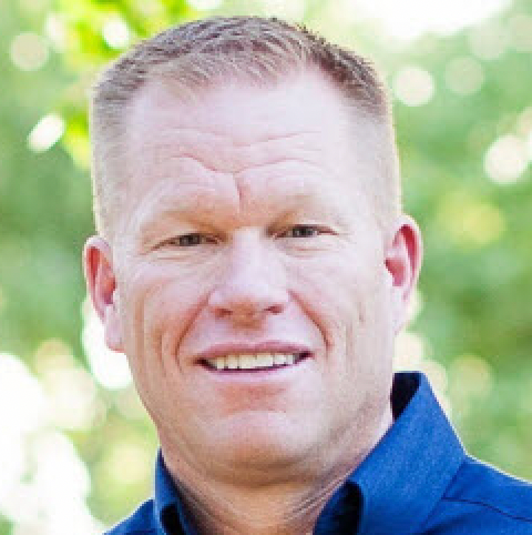 Man with short red hair wearing a blue shirt, smiling. Outside with green background.