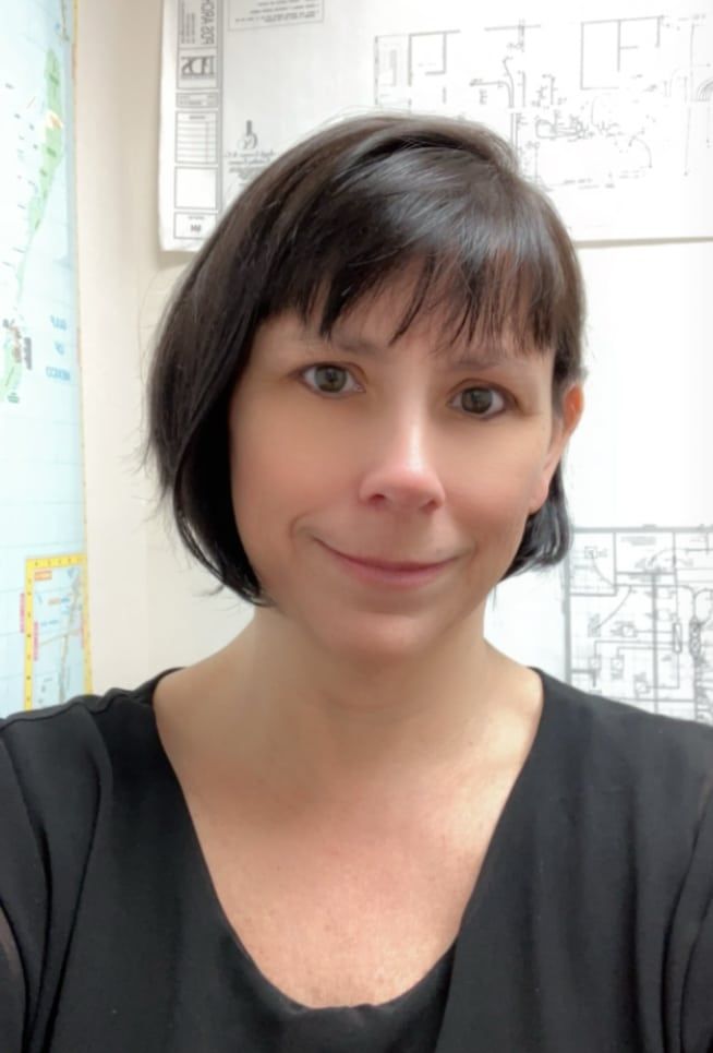 Woman with short dark hair in front of architectural plans. She is wearing a black top and smiling.