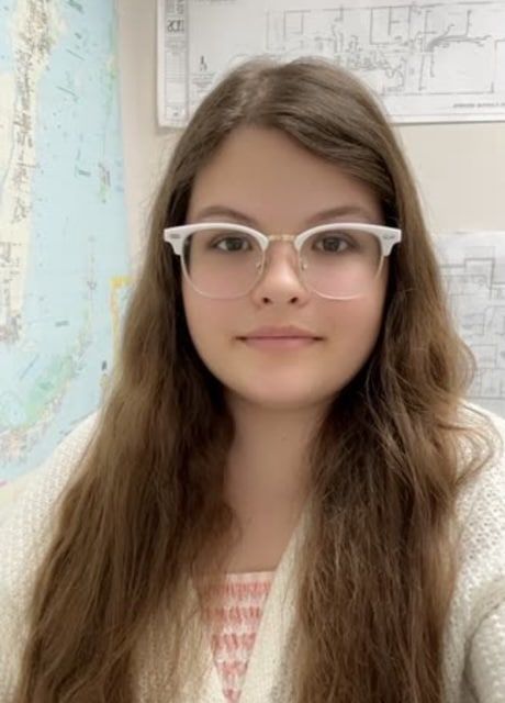 Woman with long brown hair wearing glasses, smiles at the camera in front of a world map.
